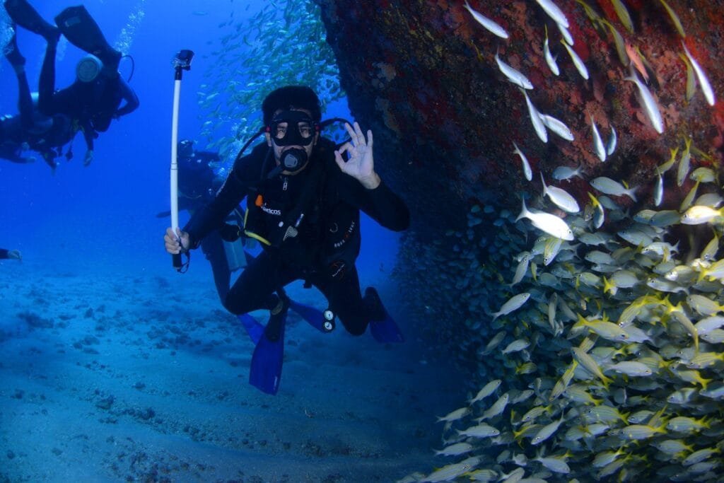 Un hombre que esta bajo el agua junto con otras personas y que practican buceo y que el esta posando y portando aletas, un traje especial para buceo y un tanque de oxígeno junto con googles y respirador